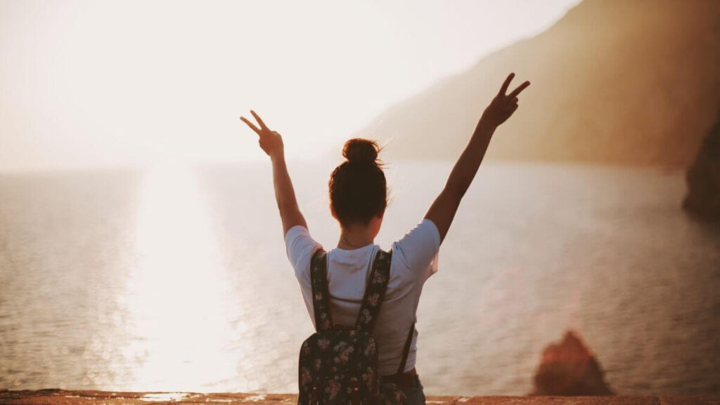 Positive Affirmationen machen happy Eine junge Frau sieht aufs meer und hebt beide Hände zum Victoryzeichen